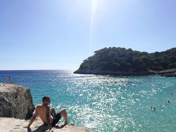 Rear view of shirtless man sitting on rock looking at sea against sky