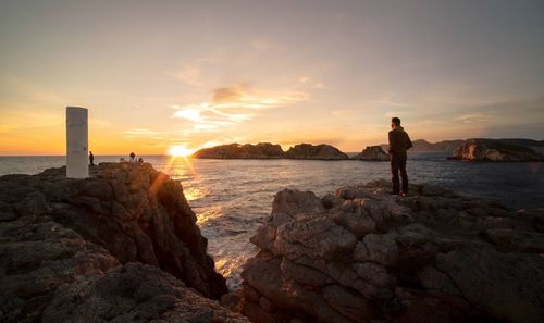 Man standing on rock by sea against sky during sunset