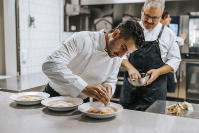 Young male chef grating cheese on dumpling plate while garnishing at countertop in kitchen