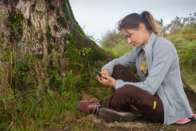 Young woman using mobile phone while sitting on field. girl with smartphone in forest.