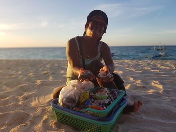 Woman holding seashell at beach against sky during sunset