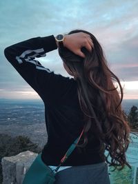 Rear view of young woman with hand in hair sitting against sky at sunset