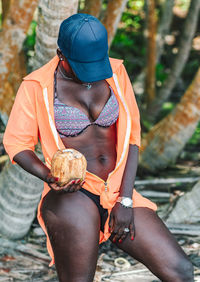 African woman with blue cap enjoys beach life and holds a newly opened coconut .