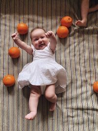 Baby girl in a white laced dress laying in a carpet playing with oranges and smiling 