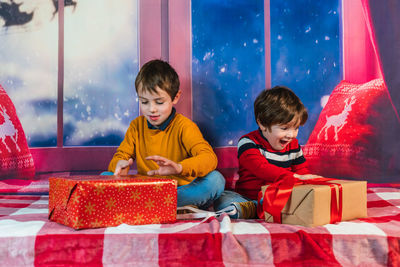 Boys sitting on checkered plaid on a background of a window and unwrapping colorful gift boxes