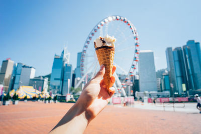 Cropped image of hand holding ice cream against ferris wheel in city