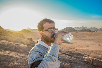 Man drinking water from bottle in desert against sky during sunset