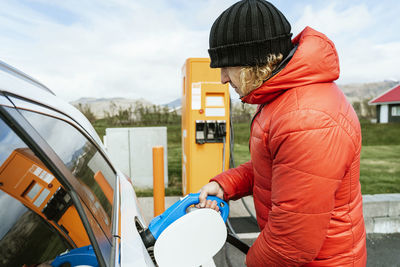 Side view of thoughtful male in warm outwear and hat standing with charging power to electric vehicle in hand at gas station in sunny day