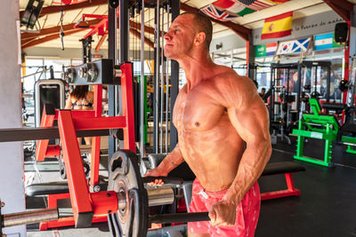 Side view of powerful male athlete with sweaty torso lifting heavy barbell plate during workout in sports club