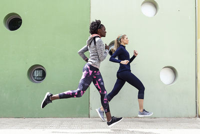 Two sporty young women running together in the city passing wall with round windows