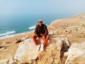 Full length of man sitting on rock at beach against sky