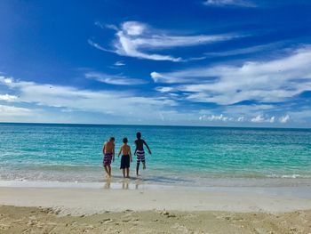 Rear view of shirtless boys standing at beach against blue sky