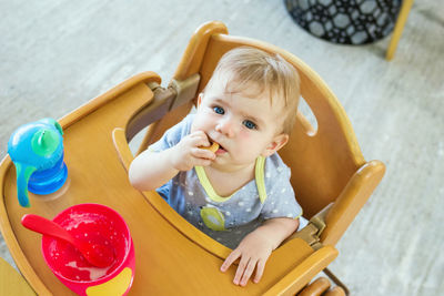 Adorable little baby girl is sitting in wooden baby chair after lunch and eating cookie.