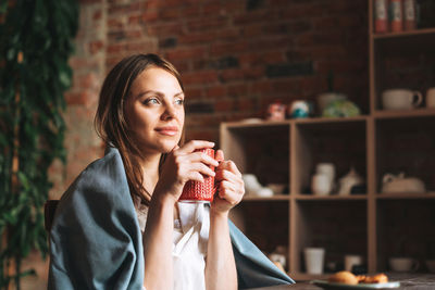 Young pretty woman in cozy grey scarf with mug of tea looks out the window and rests in her studio