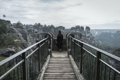 Rear view of woman standing on footbridge against sky