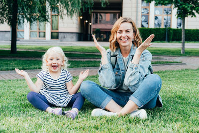 Mom and her little daughter are sitting on the lawn playing and having fun looking at the camera.