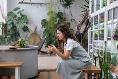 Serious young gardener woman owner holding smartphone reading media news, sitting in home garden