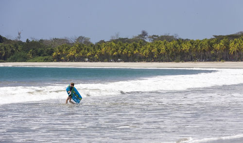 Boy with surfboard wading in sea against sky