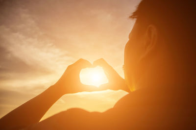 Midsection of woman holding heart shape against sky during sunset