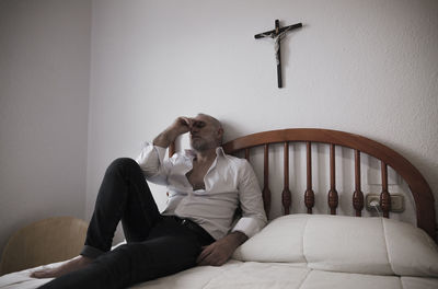 Adult man in white shirt in room lying on bed with cross on wall