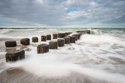 Waves splashing on wooden posts against cloudy sky