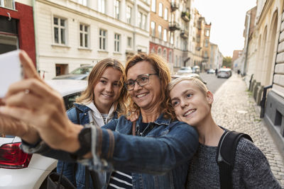 Smiling mother and children taking selfie through smart phone at roadside during weekend