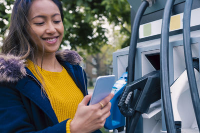 Smiling young woman using smart phone at electric vehicle charging station