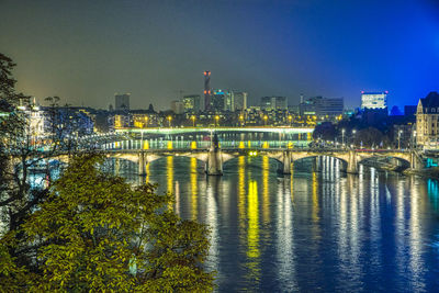 Illuminated bridge over river by buildings against sky in city