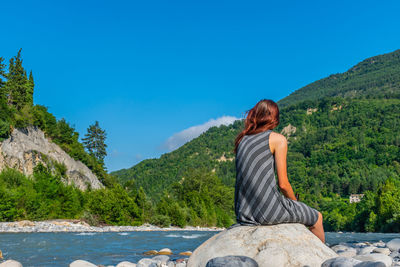 Rear view of woman sitting on mountain by lake against sky