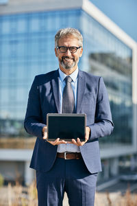 Portrait of businessman holding digital tablet while standing against office building