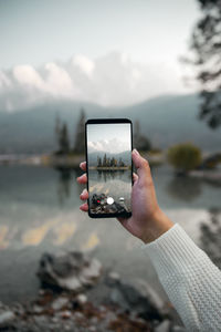 Cropped hand of woman photographing with mobile phone against lake