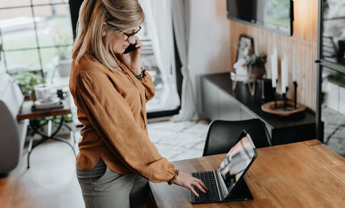Side view of businesswoman talking on phone while using laptop at home