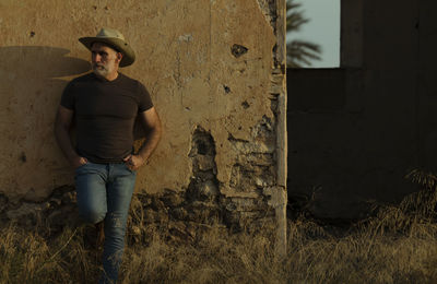 Portrait of adult man in cowboy hat and jeans against abandoned building during sunset