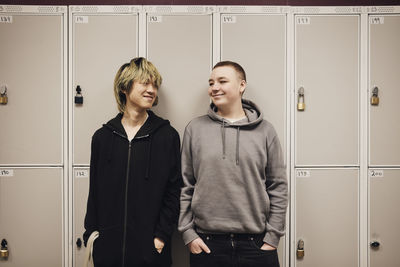 Smiling male students looking at each other while standing with hands in pockets against school locker