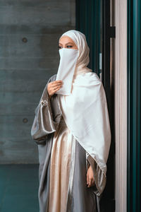 Front view portrait of veiled woman with white niqab and hijab standing against the wall
