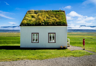 Side view of woman standing on field by house against blue sky during sunny day