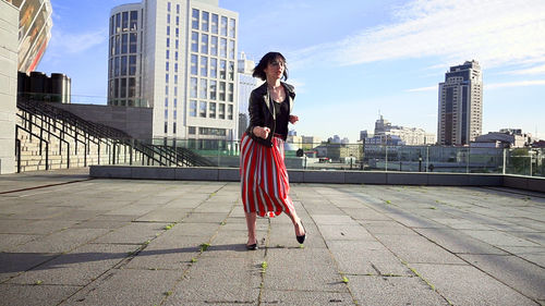 Happy woman wearing striped skirt and jacket on footpath in city