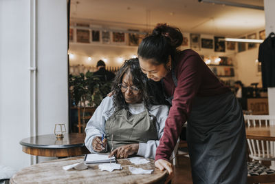 Smiling young female owner standing by colleague reading diary while sitting at table in cafe