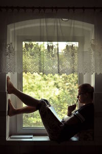 Side view of young woman studying while sitting on window sill at home