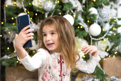 Portrait of girl photographing through mobile phone on christmas tree