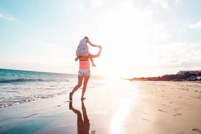 Rear view of mother carrying daughter on shoulders at beach against sky during sunset