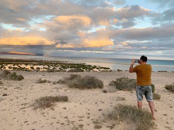 Rear view of man photographing sea on mobile phone while standing at beach against sky during sunset