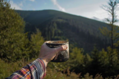 Cropped hand having drink in metal container against mountains