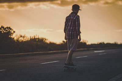 Rear view of boy skateboarding on road against sky at sunset