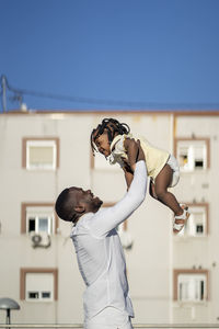 Side view of african american male standing and lifting happy little daughter while having fun on street in sunny day