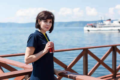 A brunette woman in a cafe on the pier is standing with a glass of juice waiting for the ship.