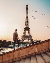 Rear view of man looking at eiffel tower while standing on railing against sky during sunset