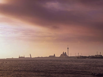 Silhouette of buildings by sea against cloudy sky