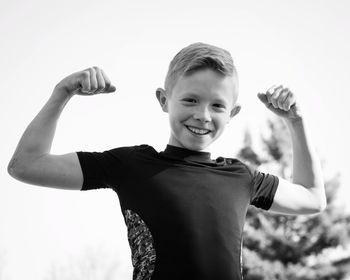 Low angle portrait of smiling boy flexing muscles while standing against clear sky