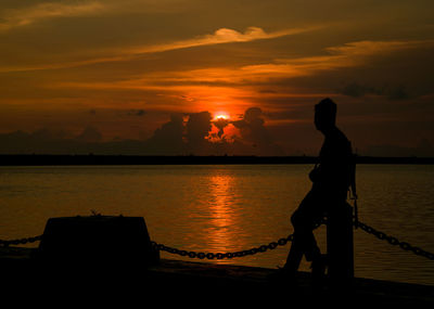 Silhouette man leaning by sea against orange sky